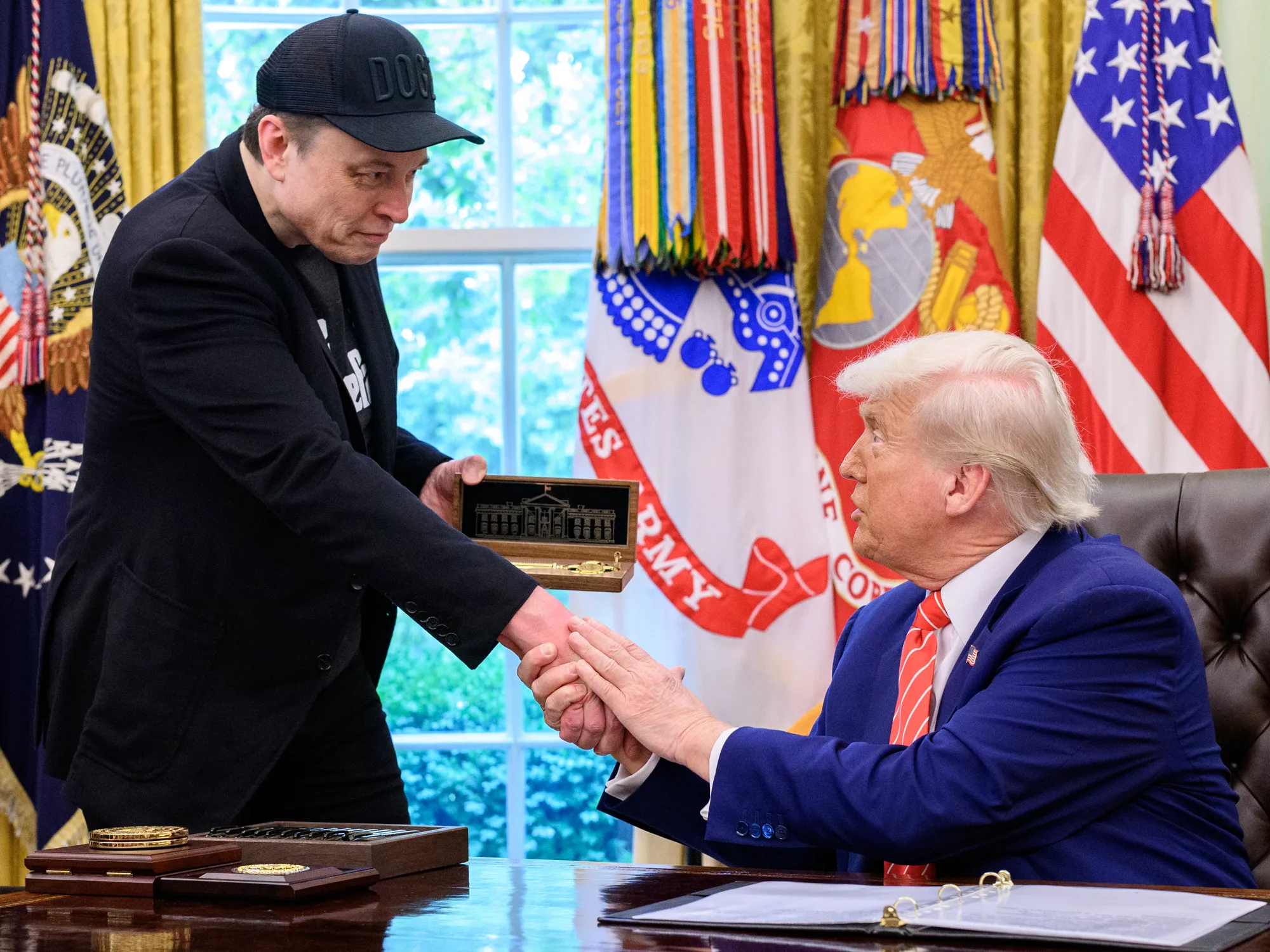 Elon Musk usando boné do DOGE cumprimenta Donald Trump no Salão Oval enquanto entrega uma caixa com maquete da Casa Branca