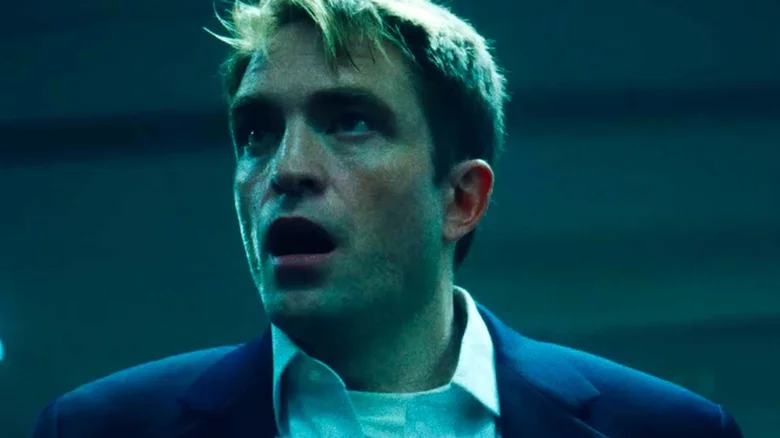 Robert Pattinson como Chris Hansen, de paletó azul-marinho e camisa branca aberta no colarinho, com a boca entreaberta e expressão de choque, sob luz azulada em cena de Primetime