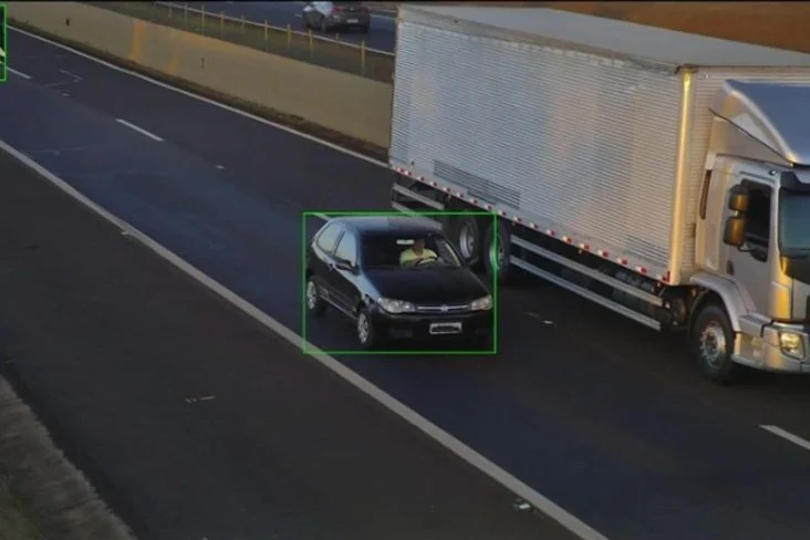 Imagem do sistema de fiscalização mostrando uma rodovia com retângulo verde de detecção desenhado em volta de um carro preto, com caminhão na faixa ao lado